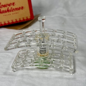 May include: A clear, vintage flower arranger with a spring mechanism. The arranger has a rectangular base and two arched, grid-like tiers. The box in the background is red and tan with the words "Flower Fashioner".