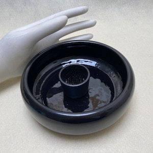 May include: A black ceramic flower frog with a round bowl and a central pin holder. The frog is designed to hold flowers in place for floral arrangements. The glossy finish reflects light, highlighting the smooth curves of the bowl.