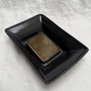May include: A black rectangular flower frog, also known as a kenzan, with a bed of brass pins. The frog sits in a black, rectangular dish with slightly raised edges. This floral arranging tool is used to hold flower stems in place.