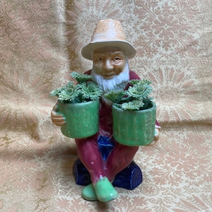 May include: A ceramic figurine of a bearded man wearing a straw hat and holding two green planter pots with green plants. The man is sitting on a blue and white log.