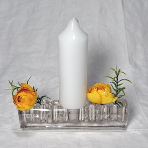 May include: A clear glass rectangular bud vase with a fluted design, holding a tall white pillar candle in the center and two bright yellow artificial flowers with green leaves placed in the side openings of the glass vessel.