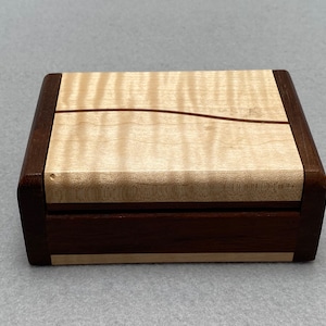 May include: A small wooden box with a light wood top and a dark wood base. The top has a wavy grain pattern and a thin dark wood inlay. The box is closed.