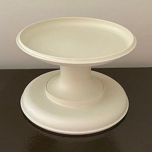 May include: A white cake stand with a round top and a wide base. The stand is made of plastic and has a smooth, glossy finish.