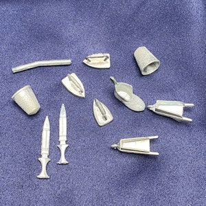 May include: A collection of miniature metal objects, including thimbles, irons, and a small chair. The objects are silver in color and have a detailed design.