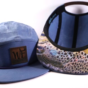 Könnte beinhalten: Eine blaue 5-Panel-Kappe mit einem braunen Leder-Patch, auf dem "WF" steht. Das Innere der Kappe ist mit einem braunen und weißen Fischdruck ausgekleidet.