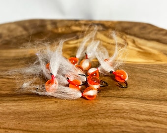 May include: A group of eight orange and white fly fishing lures with white fur tails. The lures are on a wooden surface.