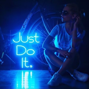 May include: A blue neon sign with the text "Just Do It."  A person wearing sunglasses and a white t-shirt sits in front of the sign.