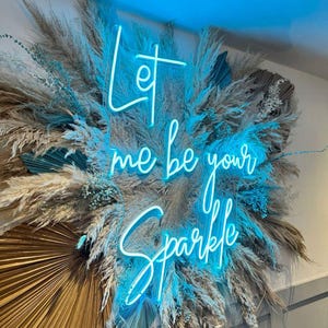 以下が含まれることがあります： 「Let me be your Sparkle」と筆記体で書かれたネオンサインの壁飾り。サインは、乾燥したパンパスグラス、青と金の扇形の葉、その他の装飾要素で囲まれています。
