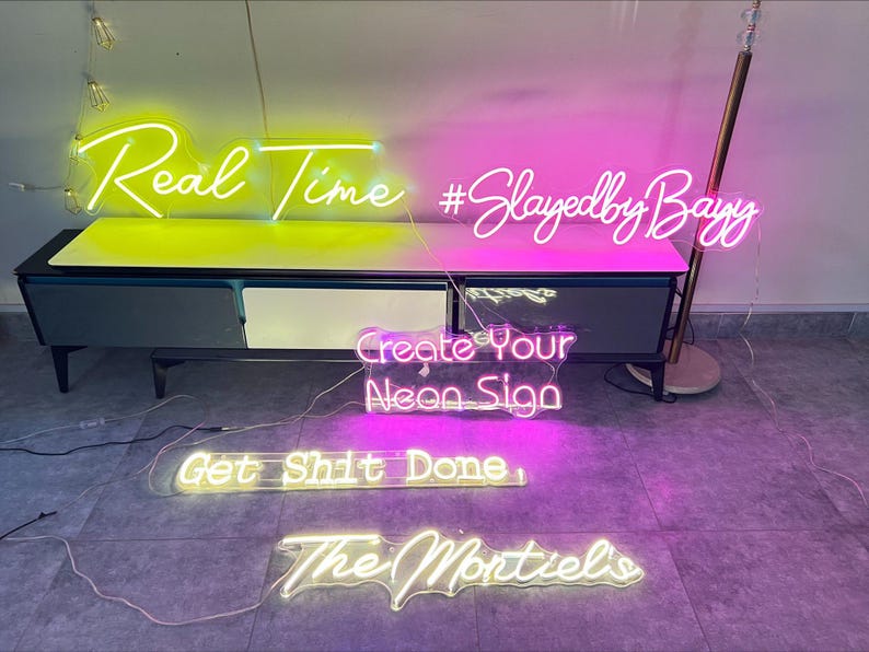 May include: Multiple neon signs featuring text in various colors and fonts. The signs display the phrases Real Time, #SlayedbyBayy, Create Your Neon Sign, Get Shit Done, and The Montiel's. These illuminated signs are arranged on and below a cabinet.