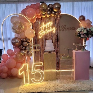 May include: A party scene with a pink and gold theme. The decor includes balloons, a cake, and neon signs that read "Happy Birthday" and the number "15". A floral arrangement and a backdrop complete the festive setting.