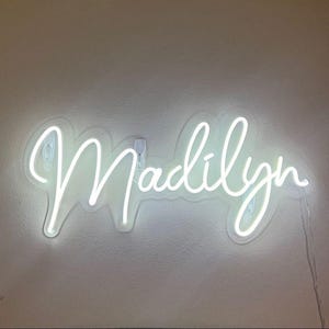 May include: A white neon sign displaying the name "Madilyn" in a cursive font. The sign is mounted on a light-colored wall, with a power cord visible.