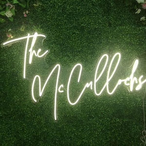 May include: A custom neon sign featuring the text The McCullochs in a white cursive script font. The sign is mounted against a dense green artificial boxwood hedge wall decorated with white and light pink silk roses and hanging floral vines.