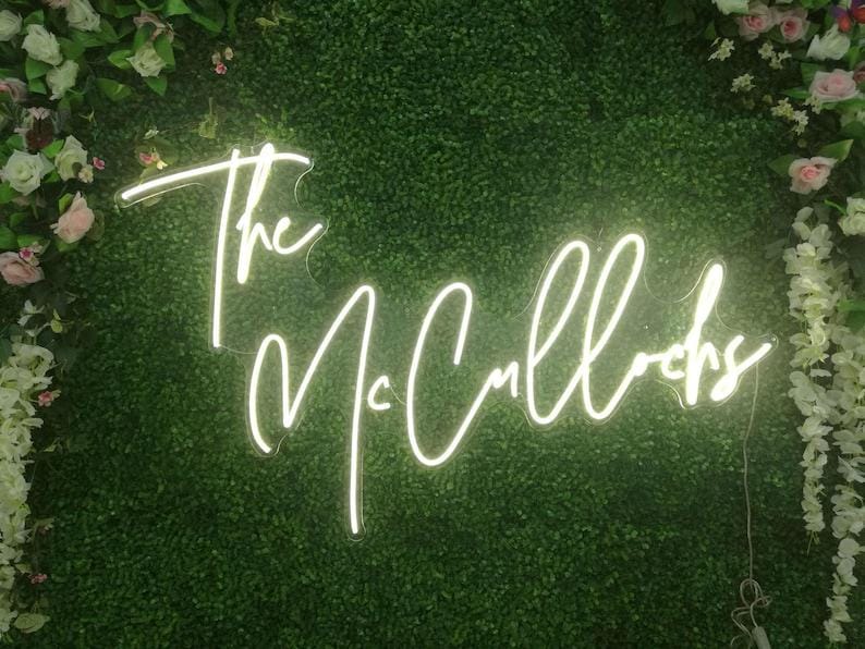 May include: A custom neon sign featuring the text The McCullochs in a white cursive script font. The sign is mounted against a dense green artificial boxwood hedge wall decorated with white and light pink silk roses and hanging floral vines.
