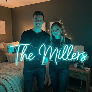 May include: A custom neon sign featuring the text The Millers in a bright teal cursive script. The sign is illuminated, casting a vibrant glow, and is held horizontally by two individuals in a bedroom setting with a bed and nightstand visible.
