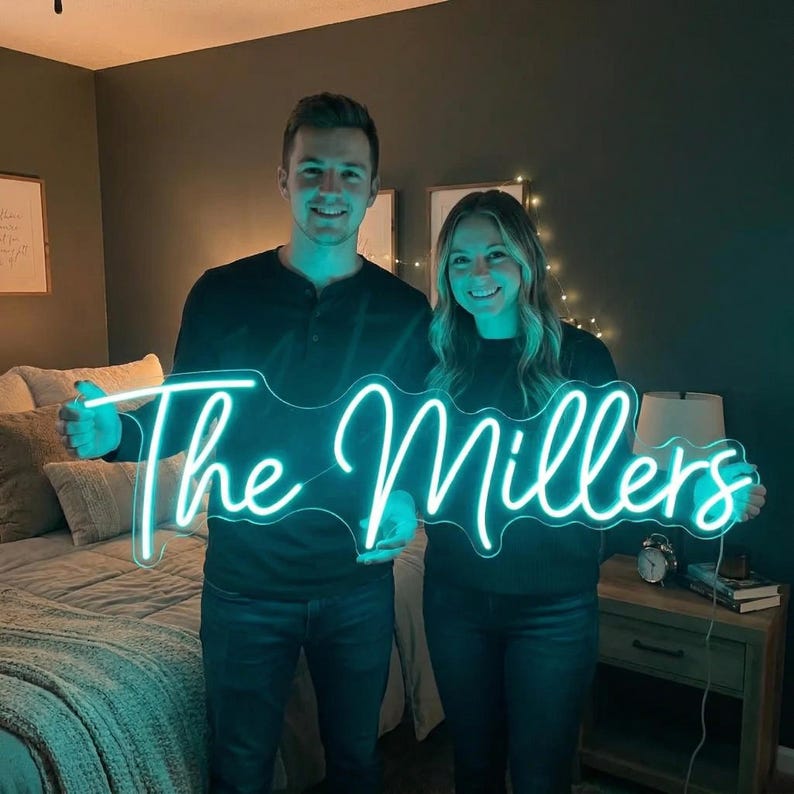 May include: A custom neon sign featuring the text The Millers in a bright teal cursive script. The sign is illuminated, casting a vibrant glow, and is held horizontally by two individuals in a bedroom setting with a bed and nightstand visible.