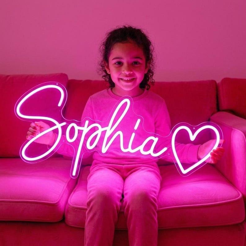 May include: A custom neon sign featuring the name Sophia in a cursive script font followed by a heart outline. The sign is constructed from white tubing that emits a bright light, creating a glowing effect against the surrounding pink environment.