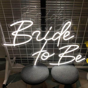 May include: A white neon sign that reads "Bride to Be" in a cursive font. The sign is mounted on a white grid background. Two gray stools are in the foreground.