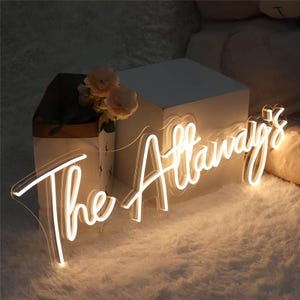 Può includere: Un cartello al neon bianco con le parole "The Attaways" in un font corsivo. Il cartello è illuminato e si trova su una superficie bianca.