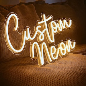 May include: A glowing neon sign featuring the words Custom Neon in a cursive script font. The sign emits a warm, bright yellow light and is mounted on a clear acrylic backing, displaying a layered effect with white and yellow illumination.