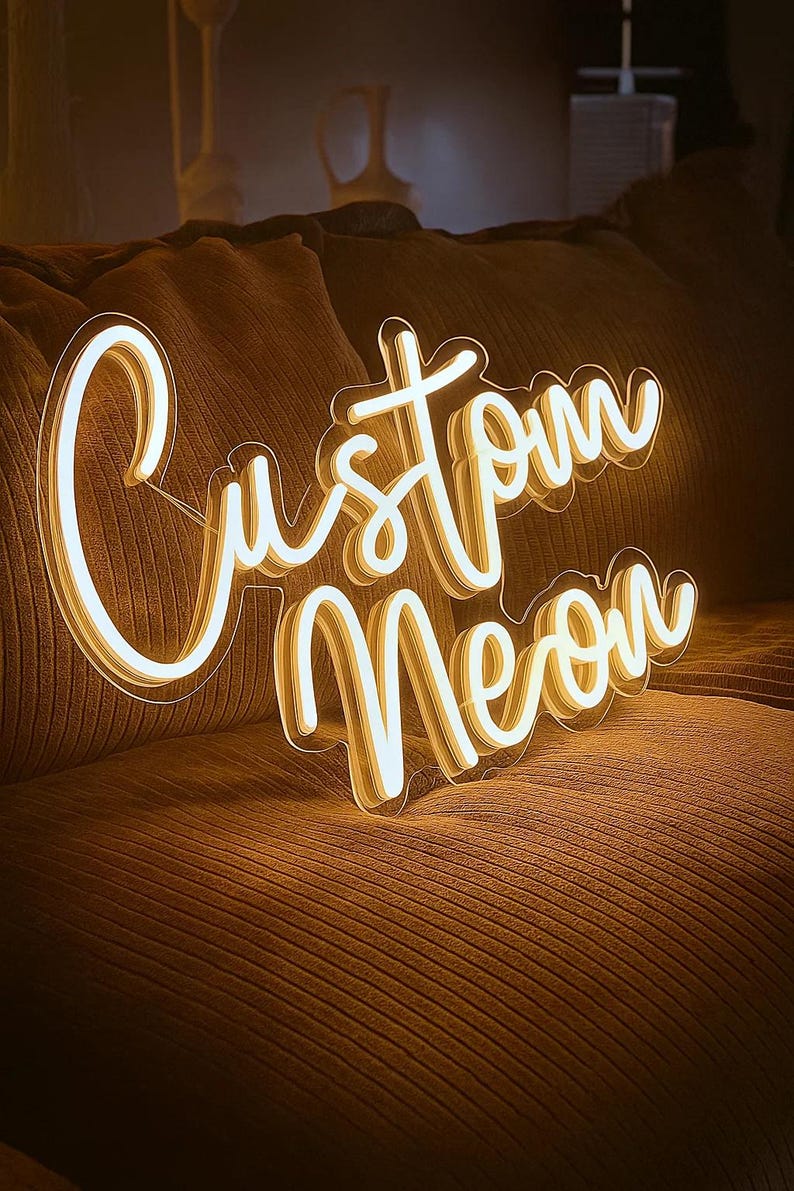 May include: A glowing neon sign featuring the words Custom Neon in a cursive script font. The sign emits a warm, bright yellow light and is mounted on a clear acrylic backing, displaying a layered effect with white and yellow illumination.