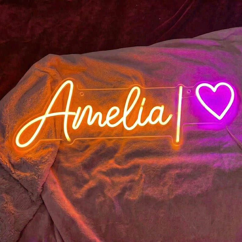 May include: A custom neon sign featuring the name Amelia in a bright orange cursive script, followed by a vertical line and a glowing pink heart symbol. The sign is mounted on a clear acrylic backing and emits a vibrant light.