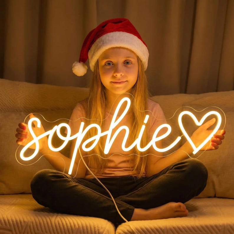 May include: A custom neon sign featuring the name Sophie in a flowing cursive script, followed by a heart symbol. The sign emits a warm, bright yellow glow and is constructed with a clear backing that follows the outline of the text and heart.