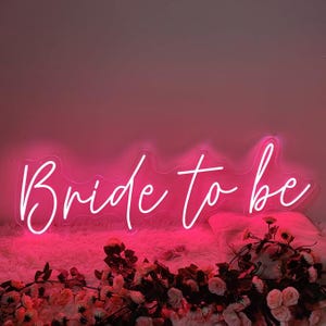 May include: A neon sign in bright pink, spelling out "Bride to be" in a cursive font. The sign is set against a soft, pink background and is surrounded by pink and white flowers, creating a celebratory atmosphere.