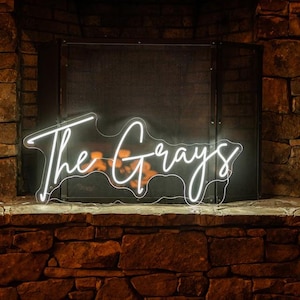 以下が含まれることがあります： 「The Grays」という文字が書かれた白いネオンサイン。サインはレンガの暖炉の前に吊り下げられている。