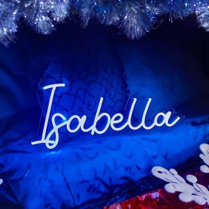 May include: A custom neon sign featuring the name Isabella in a white cursive script font. The sign is mounted on a clear acrylic backing and emits a bright white glow, positioned against a dark blue background with silver tinsel accents.