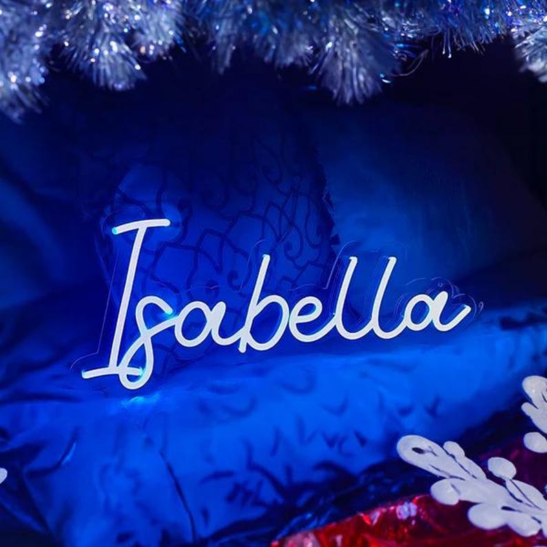 May include: A custom neon sign featuring the name Isabella in a white cursive script font. The sign is mounted on a clear acrylic backing and emits a bright white glow, positioned against a dark blue background with silver tinsel accents.