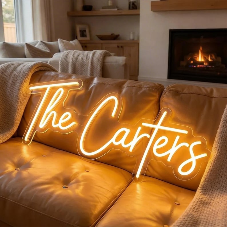 May include: A custom neon sign featuring the text The Carters in a warm, glowing yellow script font. The sign is mounted on a clear acrylic backing and emits a bright, steady light that illuminates the surrounding tan leather sofa surface.