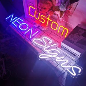 May include: Three neon signs with the words "Custom", "Neon", and "Signs" in different colors. The "Custom" sign is yellow, the "Neon" sign is purple and blue, and the "Signs" sign is white.