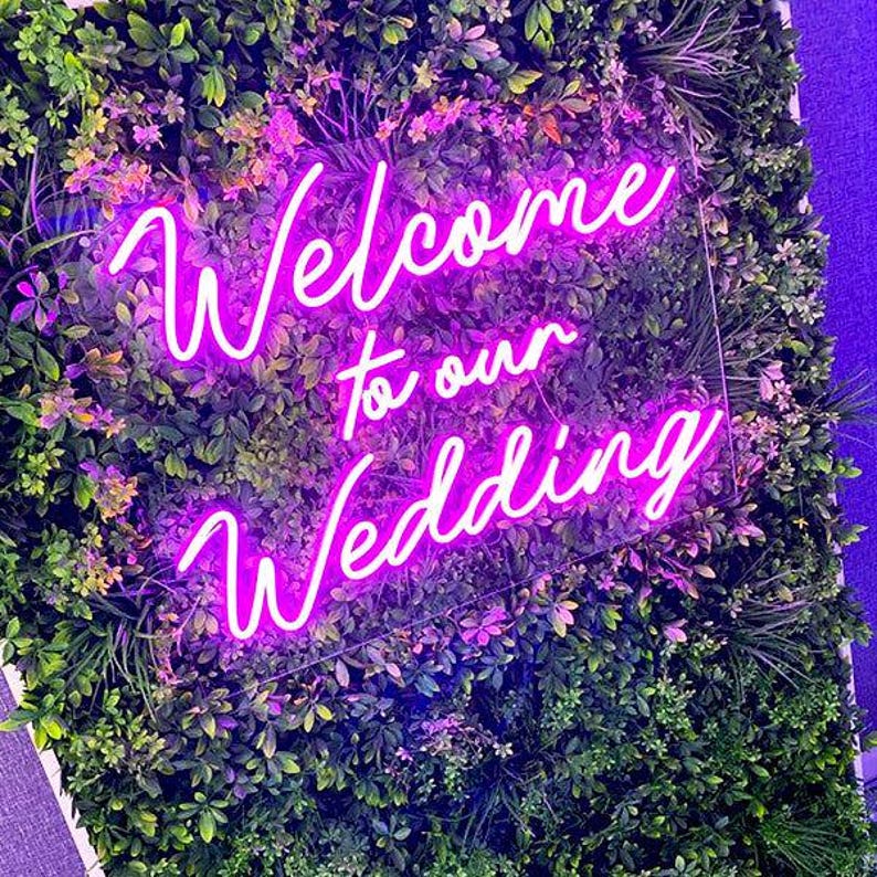 May include: A rectangular clear acrylic sign featuring bright pink neon-style lettering that reads Welcome to our Wedding. The sign is mounted directly onto a dense, lush green artificial foliage wall composed of various leaf shapes and textures.