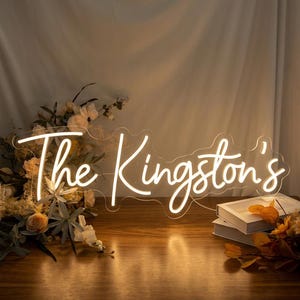 May include: A custom neon sign featuring the text The Kingston's in a glowing white script font. The sign is mounted on a clear acrylic backing and is illuminated, casting a warm light on the surrounding floral arrangement and white books.