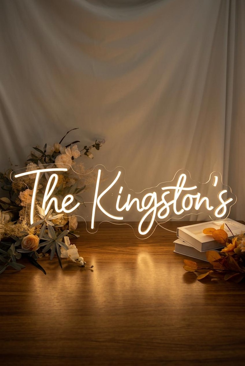 May include: A custom neon sign featuring the text The Kingston's in a glowing white script font. The sign is mounted on a clear acrylic backing and is illuminated, casting a warm light on the surrounding floral arrangement and white books.
