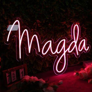 May include: A neon sign in a cursive font spelling the name "Magda" in bright pink. The sign is illuminated against a dark background with greenery. A calendar shows "SEP 1" and flowers are in the foreground.