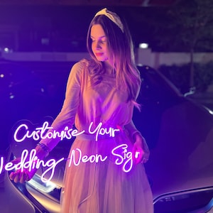 May include: A woman holds a custom neon sign that says "Customise Your Wedding Neon Sign" in white lettering with a purple glow. The sign is held in front of a silver car.