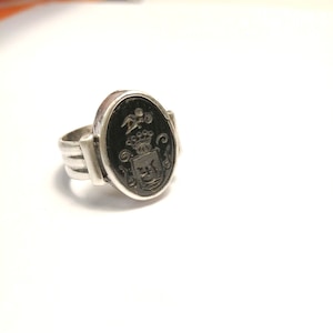 May include: A silver ring with an oval, black stone featuring a detailed crest design. The ring has a thick band with multiple parallel lines, and the stone is set in a silver bezel.