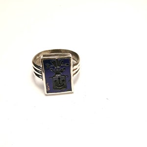 May include: A silver ring with a rectangular, dark blue face. The face features a black crest design with a crown, arrow, and anchor. The ring has a triple band design.
