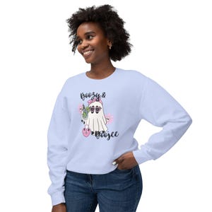 May include: A light blue long-sleeve sweatshirt featuring a graphic of a ghost wearing a pink headband and heart-shaped sunglasses, holding a cocktail and a heart-shaped purse, with the text "Boozy & Boojee" printed in black script lettering.