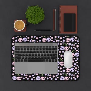 May include: A black mousepad with a repeating pattern of skulls, cherries, hearts, and flowers in pink, purple, and white. A laptop, white mouse, and other office supplies are on the desk. A cup of coffee and a small plant are also visible.