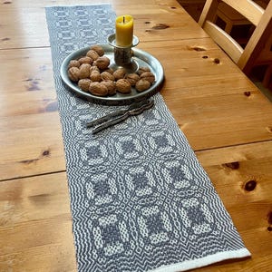 Könnte beinhalten: Ein gewebter Tischläufer in Grau und Weiß mit geometrischem Muster. Der Läufer liegt auf einem Holztisch mit einer Kerze und einem Teller mit Walnüssen.