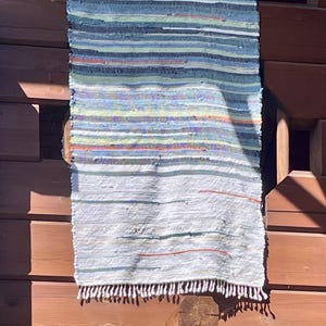 Könnte beinhalten: Ein rechteckiger, handgewebter Teppich mit einem Streifenmuster in Blau-, Grün-, Orange- und Weißtönen. Der Teppich hat einen Fransenrand und hängt an einer Holzkonstruktion.