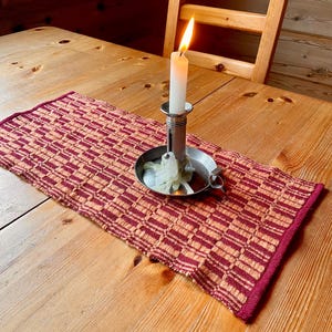 Könnte beinhalten: Eine brennende weiße Kerze in einem silbernen Kerzenhalter steht auf einem Holztisch. Unter der Kerze liegt ein gewebter Tischläufer mit einem roten und beigen geometrischen Muster. Im Hintergrund steht ein Holzstuhl.