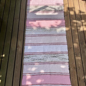 Könnte beinhalten: Ein rechteckiger gewebter Teppich mit abwechselnden horizontalen Streifen in Rosa, Weiß und Grau. Der Teppich hat an beiden Enden einen Fransenrand. Der Teppich besteht aus einem gewebten Material.