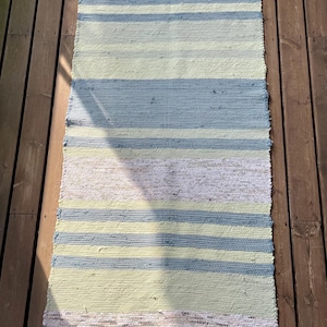 Könnte beinhalten: Ein rechteckiger, handgewebter Teppich mit horizontalen Streifen in Hellblau, Hellgelb und Weiß. Der Teppich hat einen Fransenrand und wird auf einer Holzterrasse präsentiert. Dieser Teppich ist für den Innen- und Außenbereich geeignet.