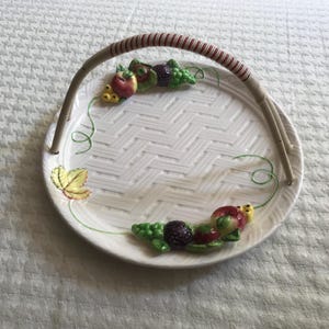 May include: White ceramic serving tray with a woven texture and a brown and red striped handle. The tray is decorated with colourful fruit and leaf accents, including apples, grapes, and blackberries. A decorative piece for serving food.