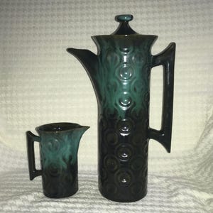 May include: A vintage teal and black ceramic coffee pot and creamer set. The coffee pot has a tall, slender design with a lid and a handle. The creamer is small with a handle. Both pieces have a circular pattern and a glossy finish.