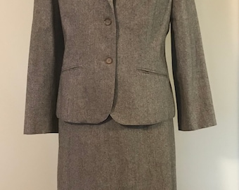 May include: A two-piece suit in a textured taupe fabric, featuring a blazer with notched lapels, two front buttons, and two welt pockets, paired with a matching knee-length skirt. A silver-toned metallic brooch is pinned to the left lapel.
