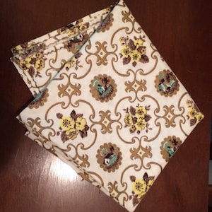 May include: Folded vintage fabric featuring a cream backdrop and a repeating design. The pattern incorporates yellow flowers, brown scrollwork, and circular medallions with a small house illustration. The fabric is folded on a dark brown surface.